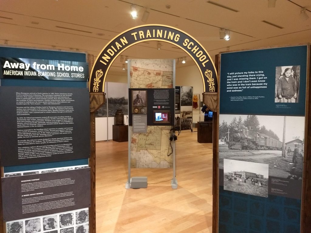 Image of the opening of an exhibition, featuring image and text panels and an arch with the words "Indian Training School."