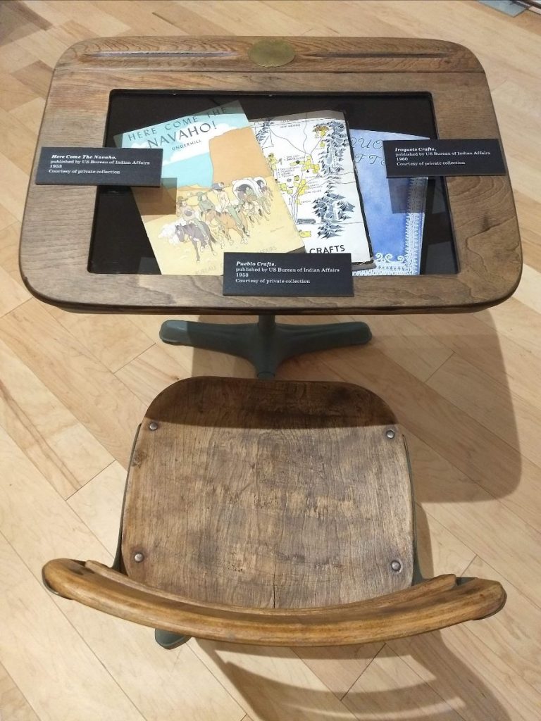 Looking down at a small wooden classroom desk and chair. The desk has a clear pane set in its lid, under which are three books.