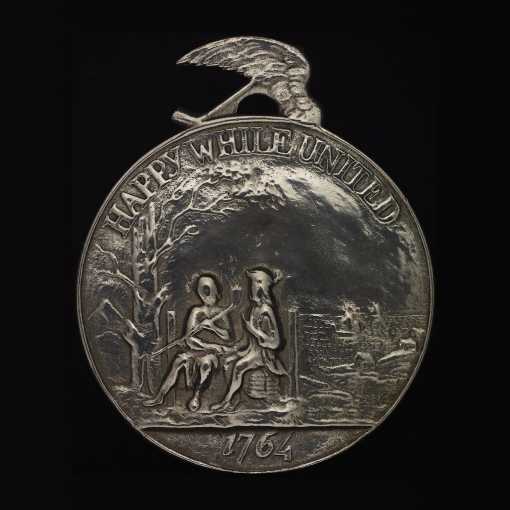 A silver medal with a relief image of two seated people with a pipe under a tree in the foreground and a landscape featuring houses and vessels on water in the background. Text at the top reads "HAPPY WHILE UNITED" and at the bottom is the date "1764".